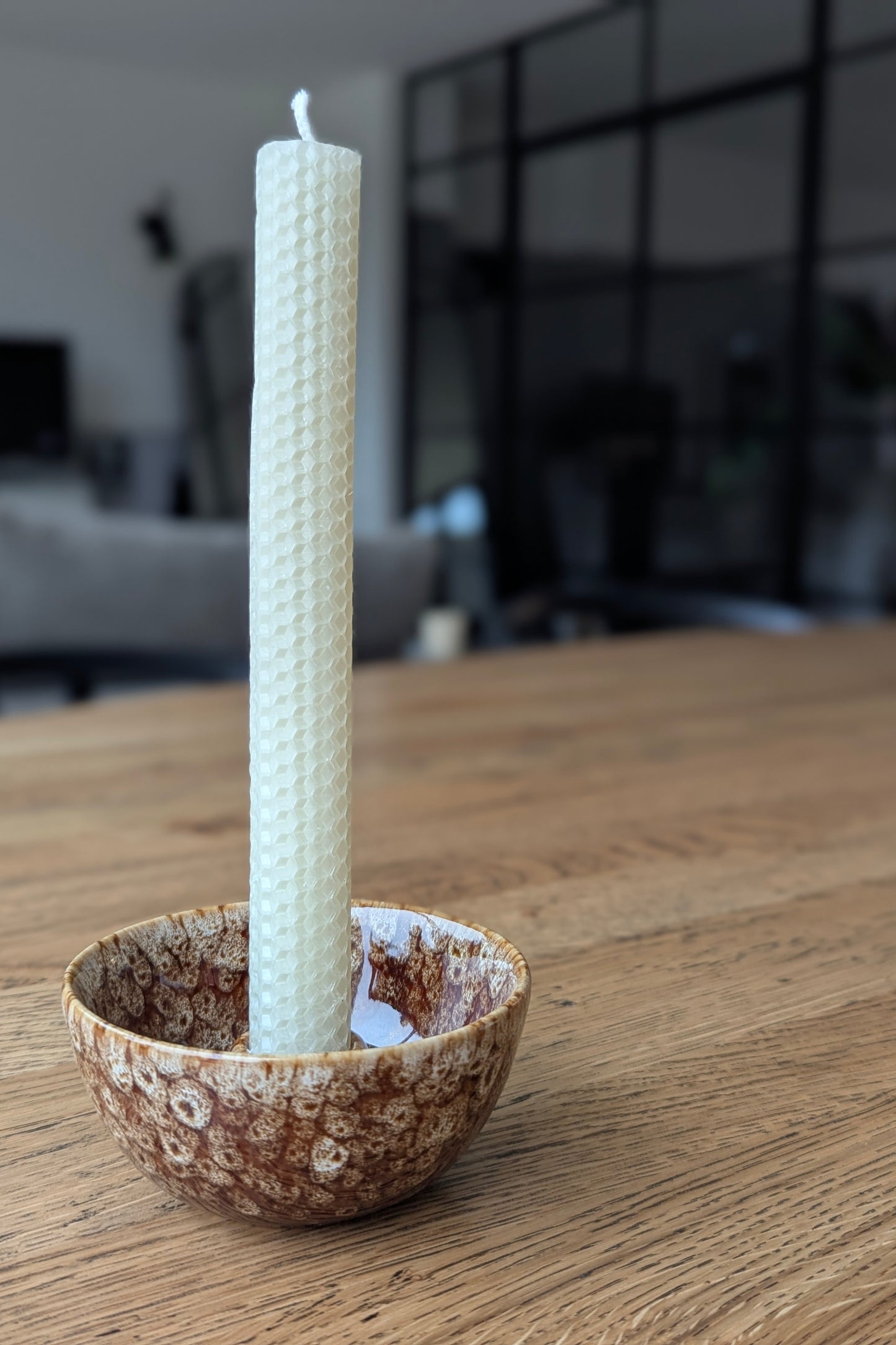 White candle in a textured ceramic holder on a wooden surface with a blurred indoor background