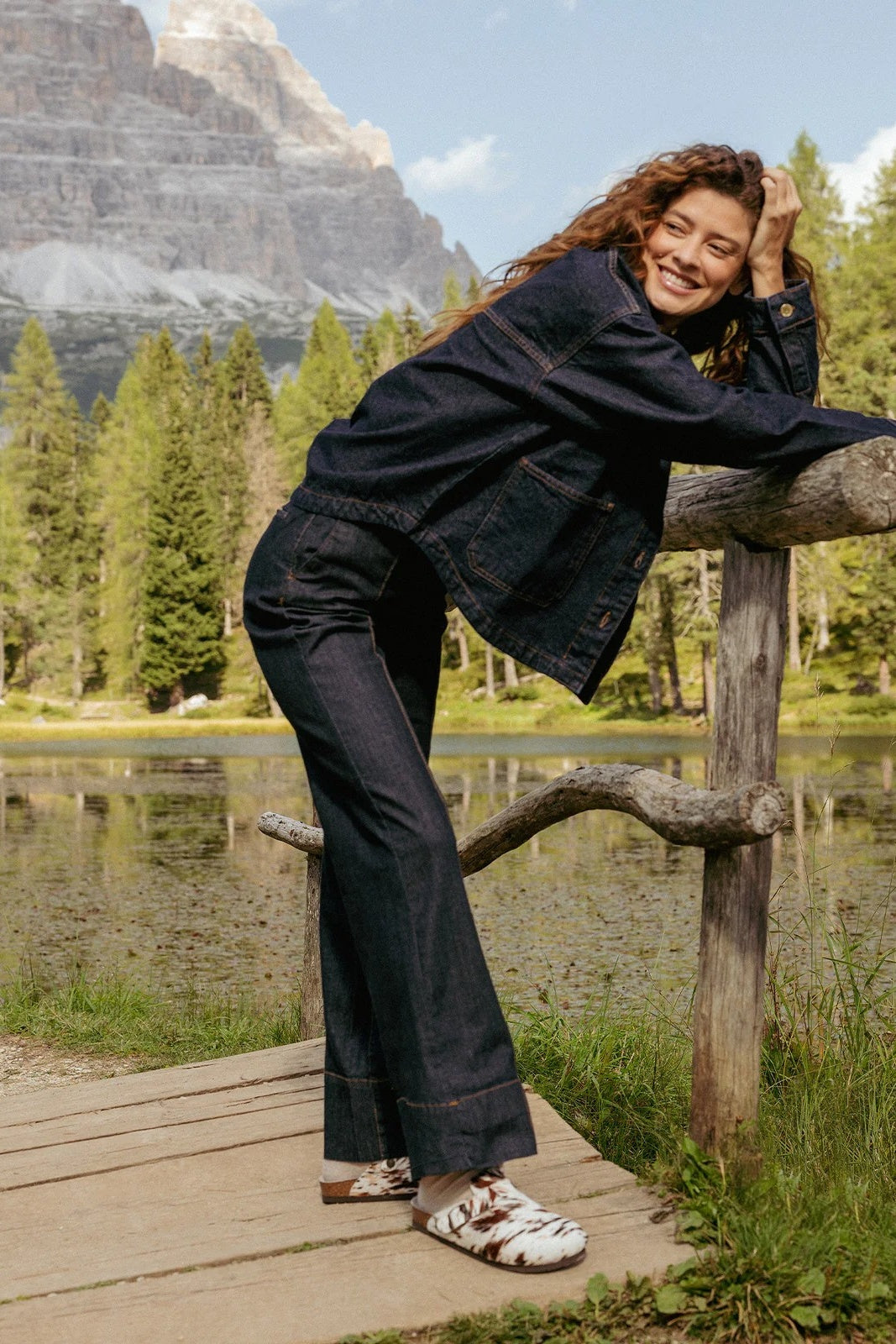 Woman in a black outfit leaning on a wooden fence with a mountain and lake in the background