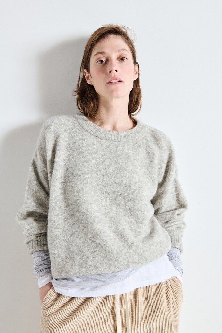 Woman wearing a light grey sweater over a white shirt with beige pants against a plain background