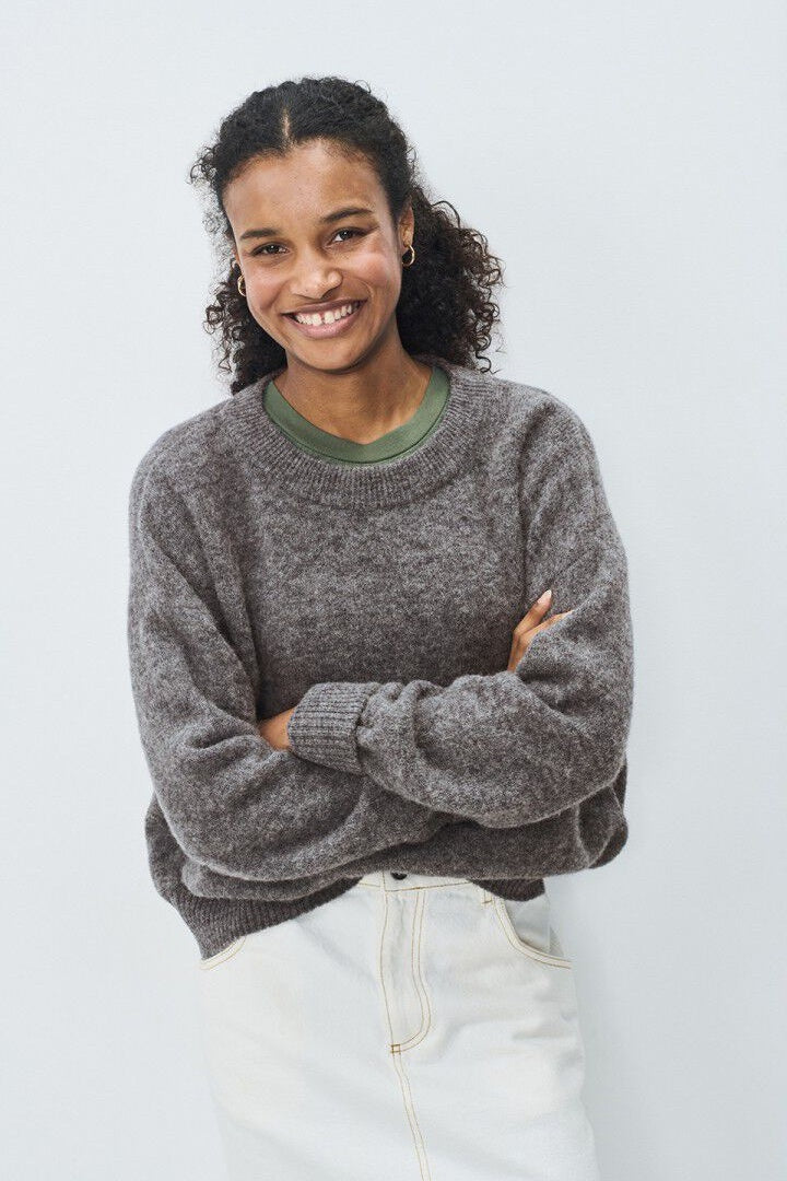 Woman wearing a grey sweater and white pants against a light background