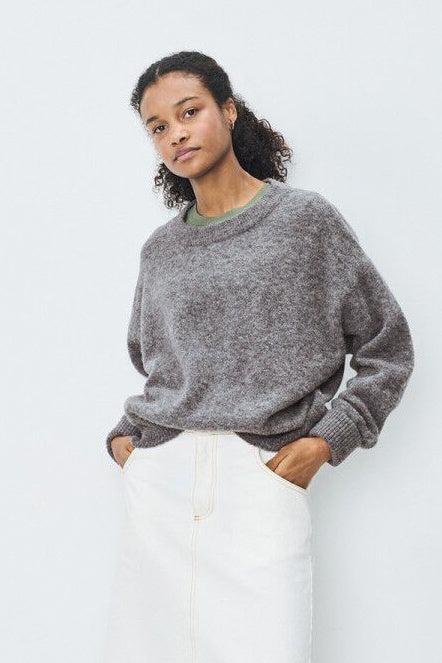 Woman wearing a grey sweater and white pants against a plain background