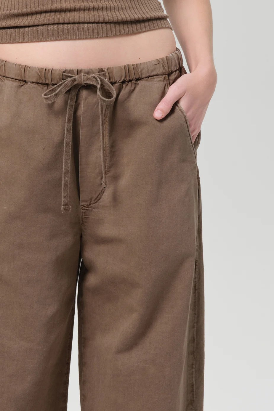 A close-up of a person wearing brown wide-leg drawstring pants, showing the waistband, side pocket, and hand in the pocket, paired with a matching ribbed top against a light gray background.