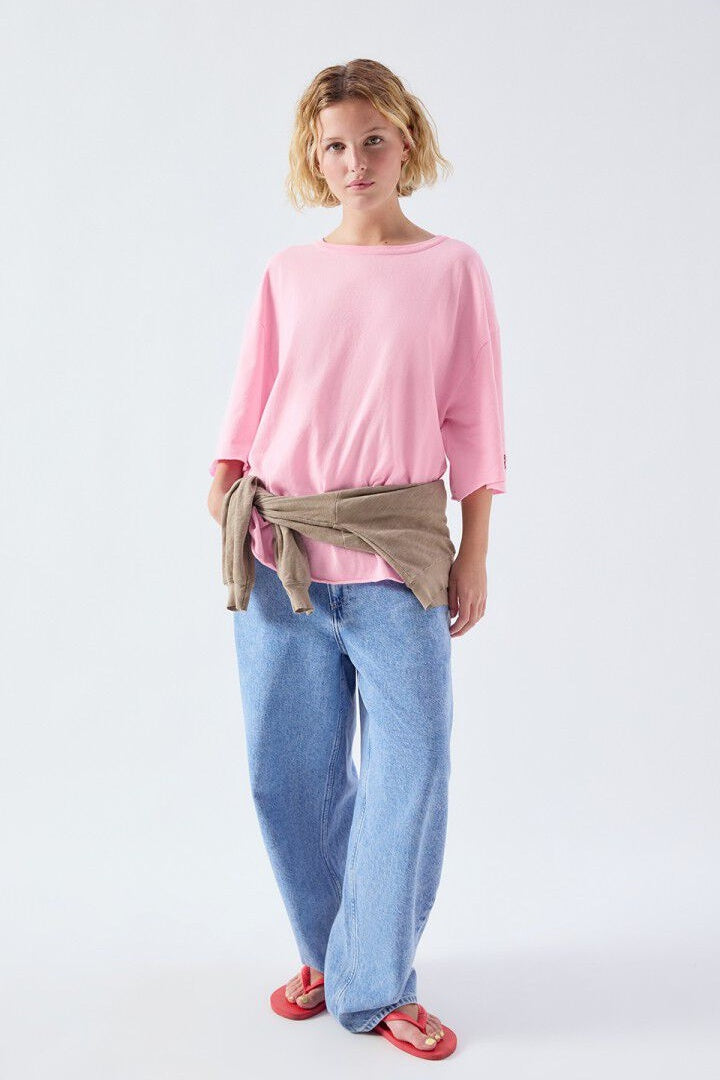 Woman wearing a pink sweater, beige jacket, blue jeans, and red sandals on a white background