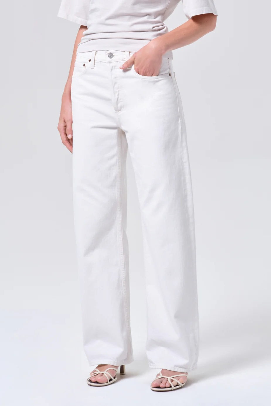 White wide-leg jeans worn by a model, paired with a white t-shirt and beige strappy sandals, shown from the waist down on a light gray studio background.