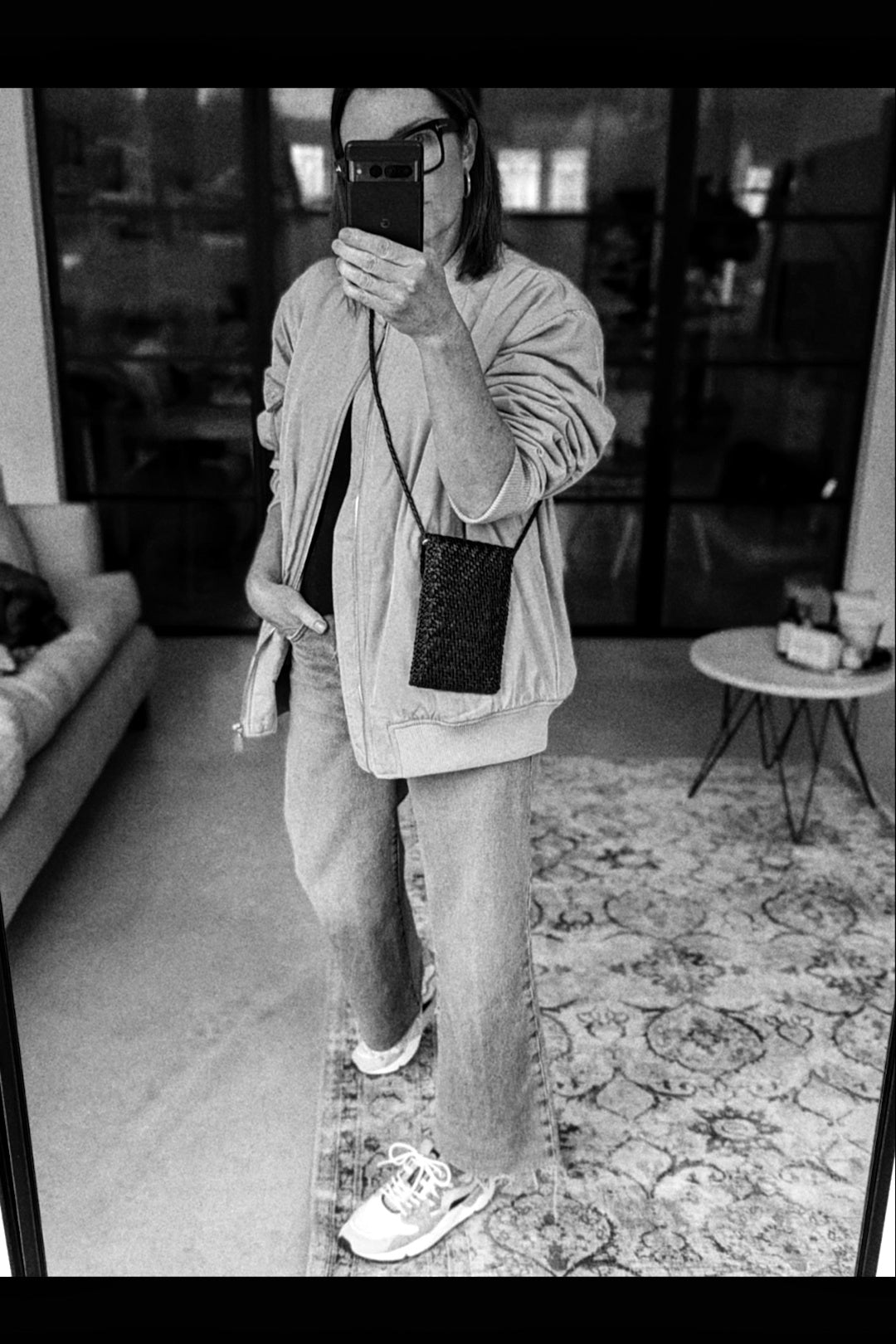 Person taking a mirror selfie in a living room wearing a light jacket, dark pants, and white sneakers.