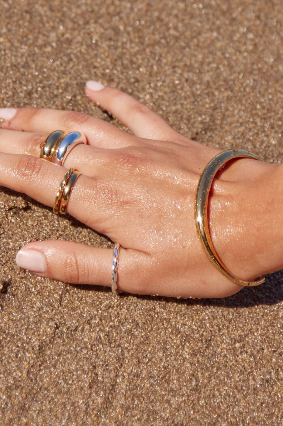 Hand with multiple gold rings and a bracelet on a sandy background