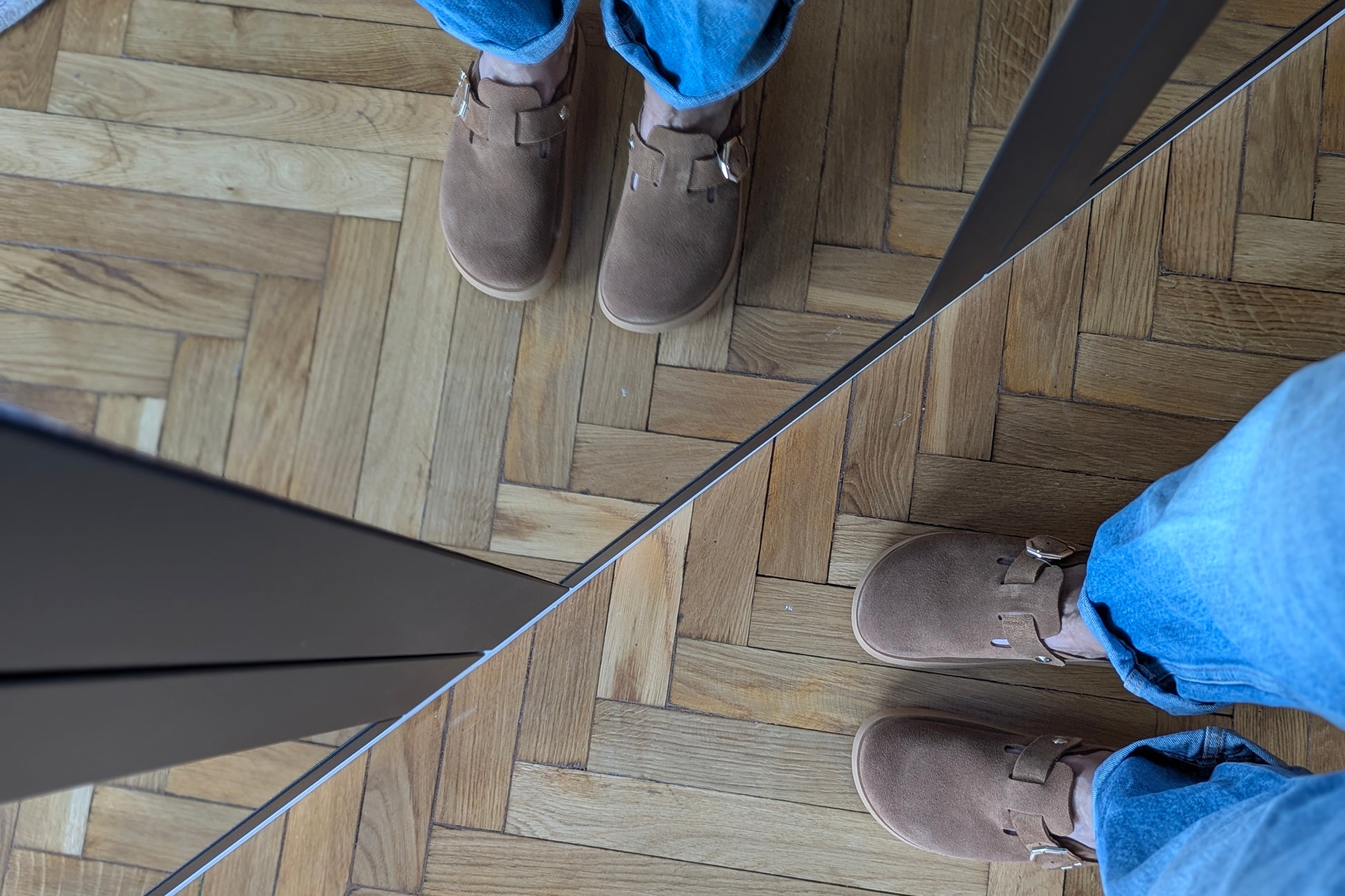 Person wearing brown clog shoes and blue jeans standing on a wooden floor.