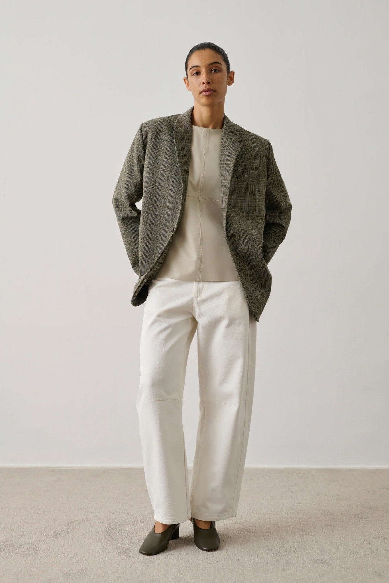 Person wearing a beige jacket, beige sweater, and white pants against a plain background