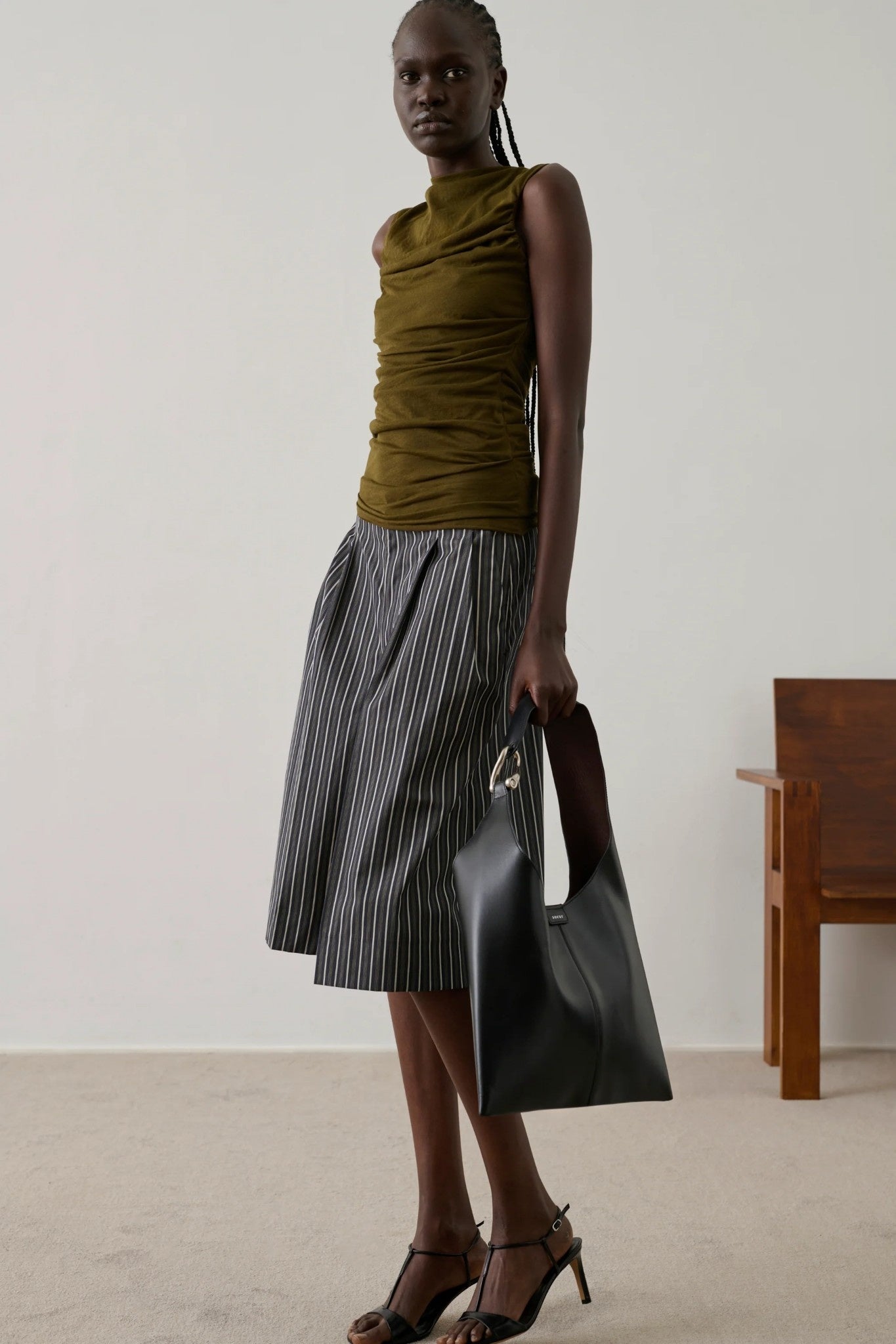 Woman in bronze top and striped skirt holding a black handbag against a plain background