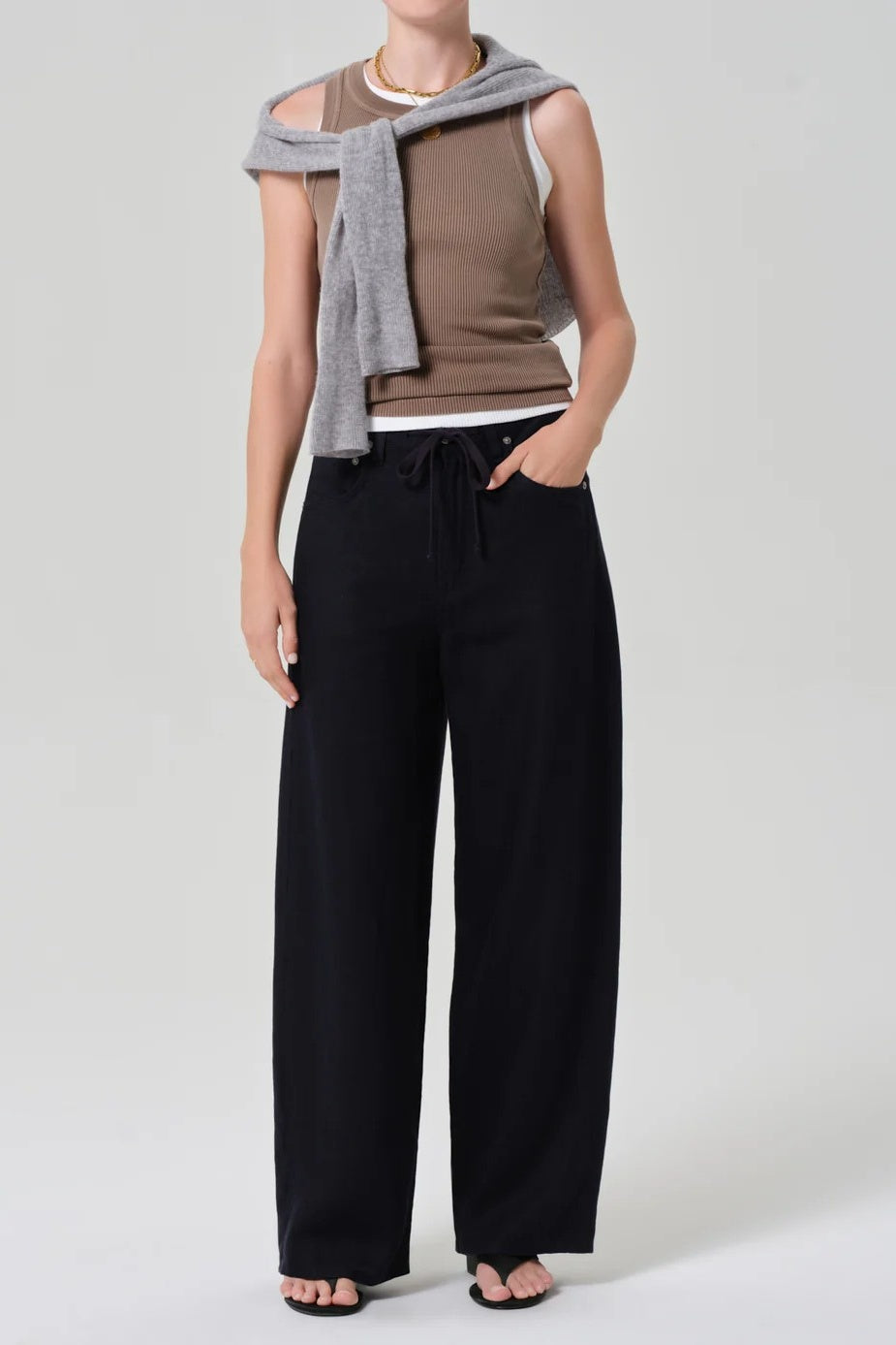 A model wearing black wide-leg drawstring pants, a brown sleeveless top, and a gray knit sweater draped over her shoulders, photographed against a light gray studio background.