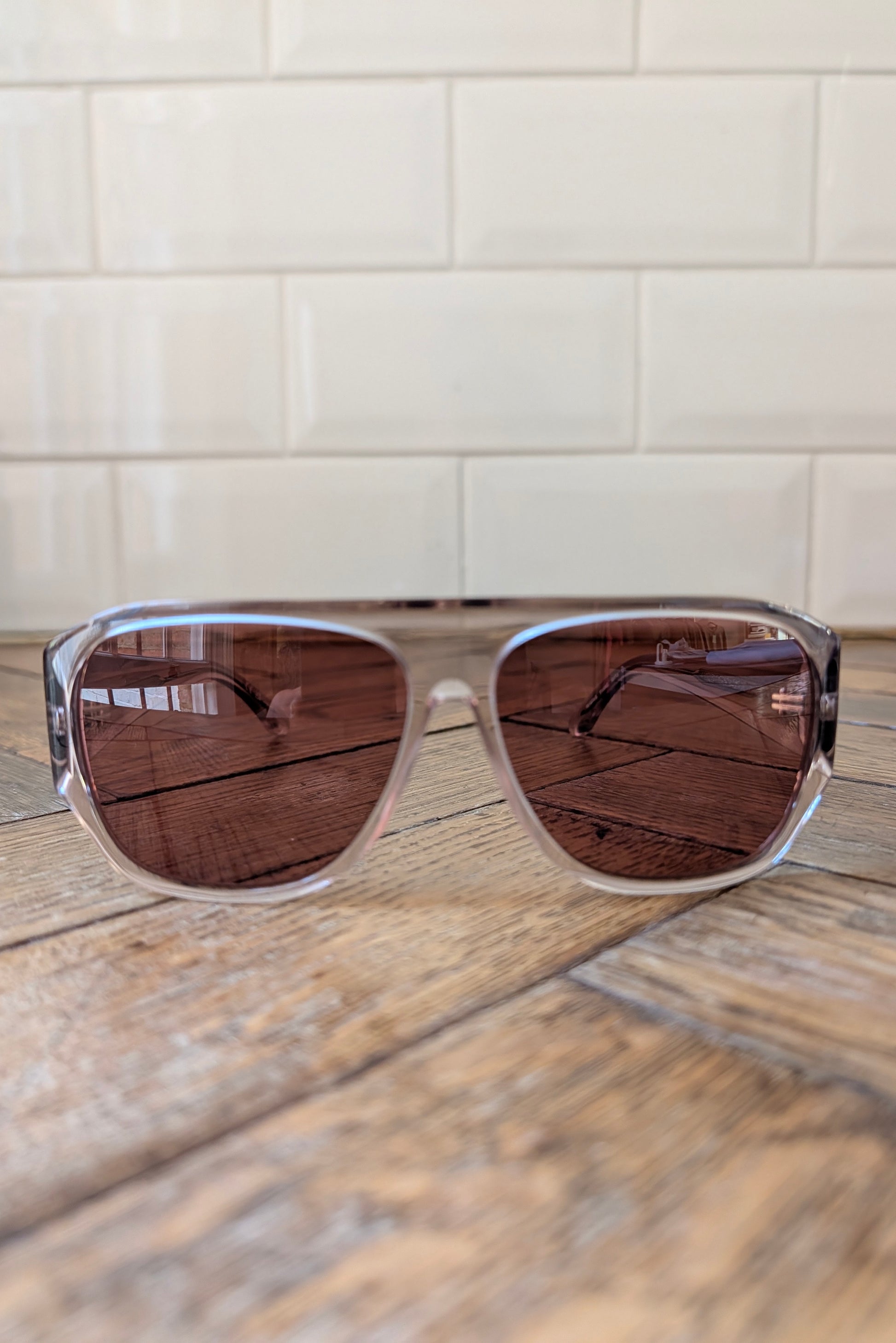 A pair of square aviator-style sunglasses with a translucent light gray frame and dark brown tinted lenses, resting on a rustic wooden surface with a white subway tile wall in the background.