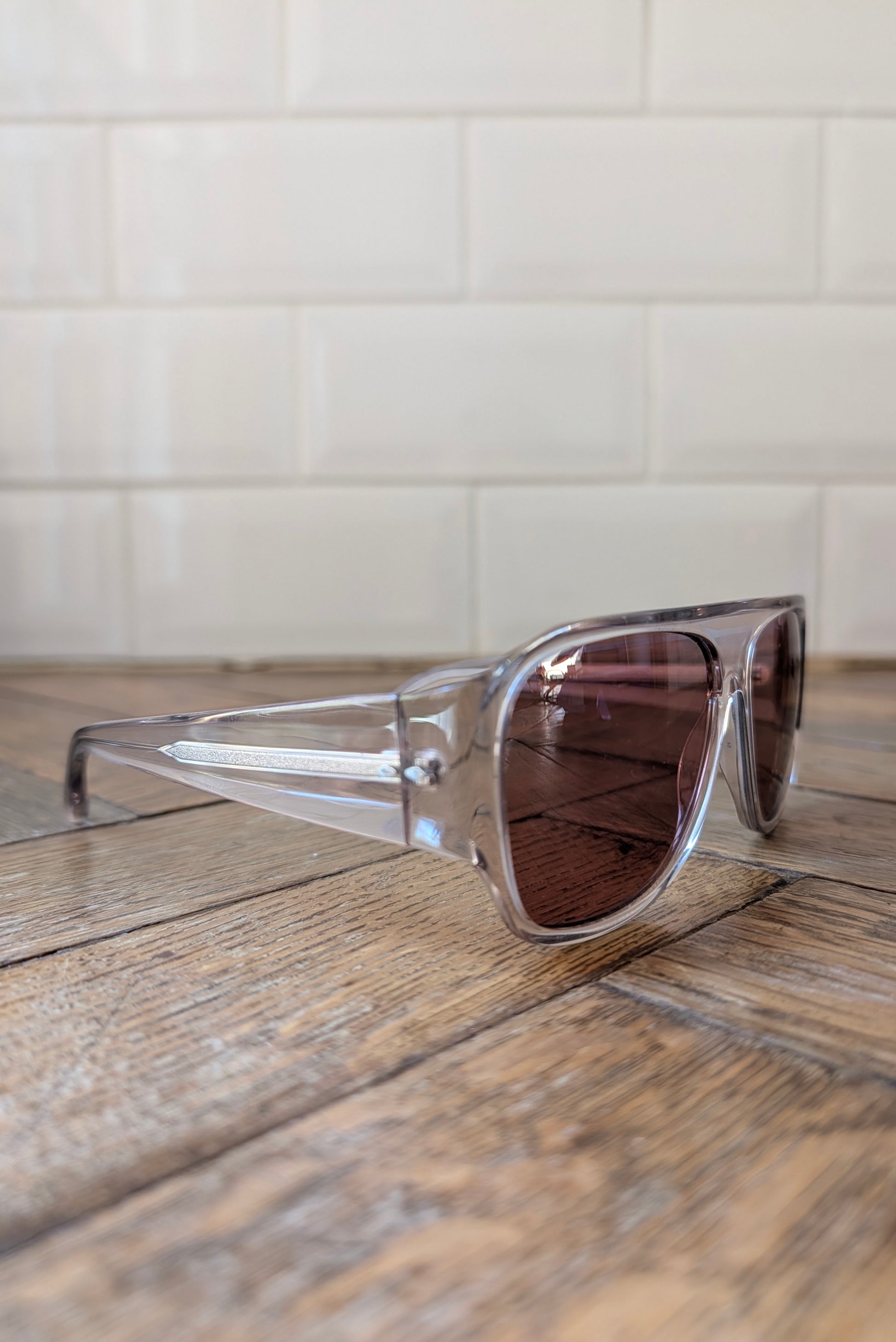Clear acetate square sunglasses with brown tinted lenses resting on a rustic wooden surface against a white subway tile wall.