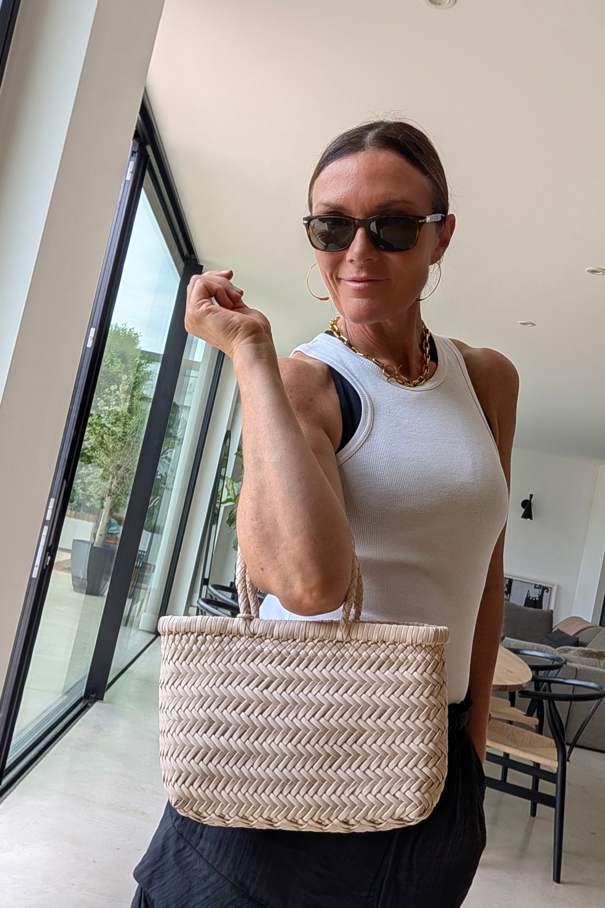 Woman holding a woven bag indoors, wearing sunglasses and a white top.