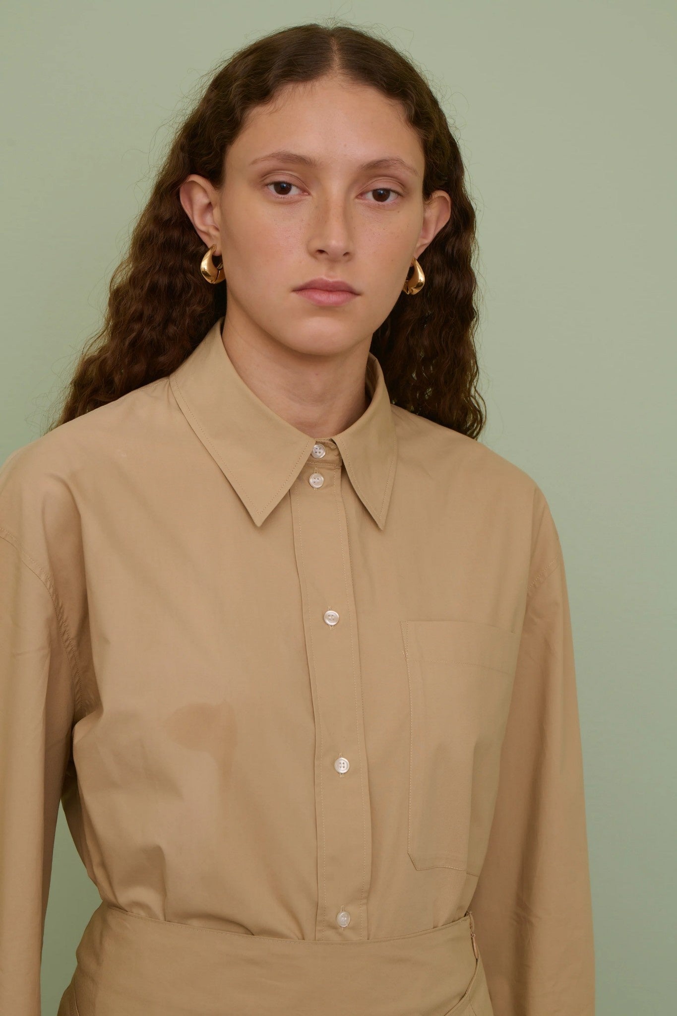 Woman wearing a beige shirt and gold earrings against a light green background