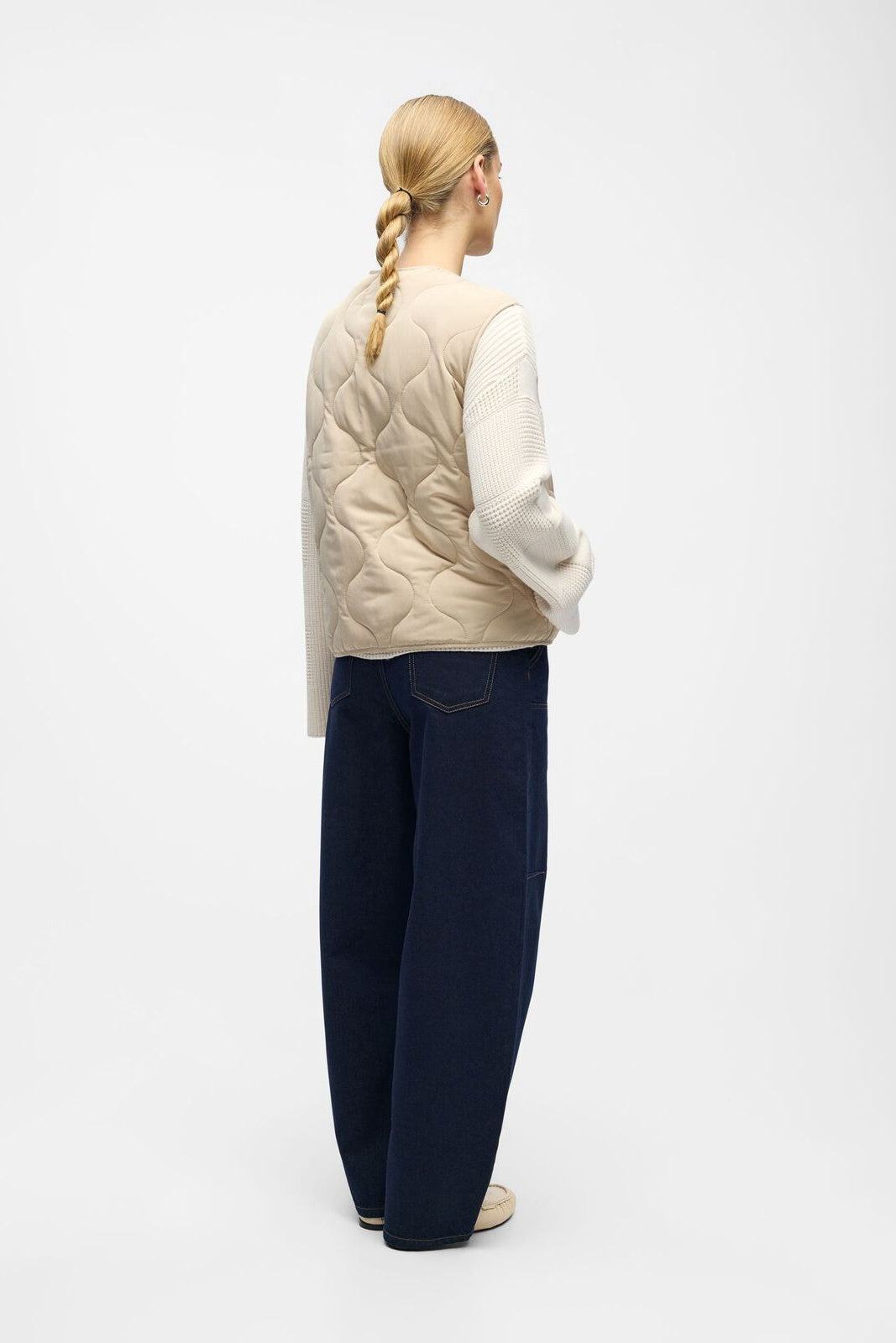 Person wearing a beige quilted vest, white sweater, and navy pants on a white background