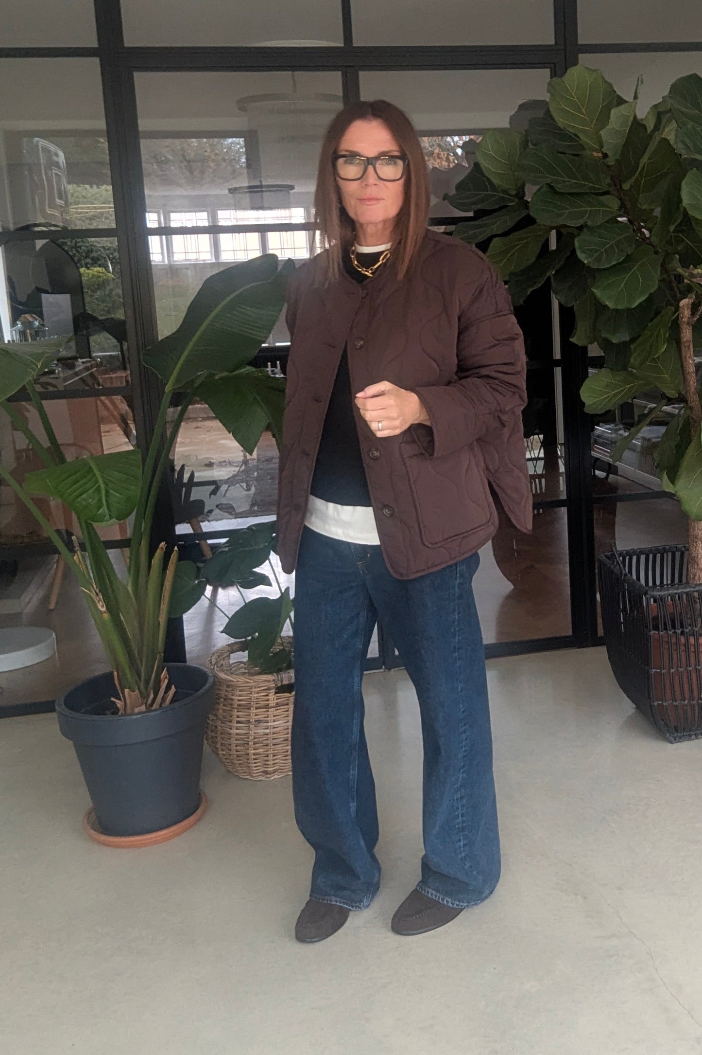 Woman wearing a brown coat and blue jeans standing indoors with plants and furniture in the background