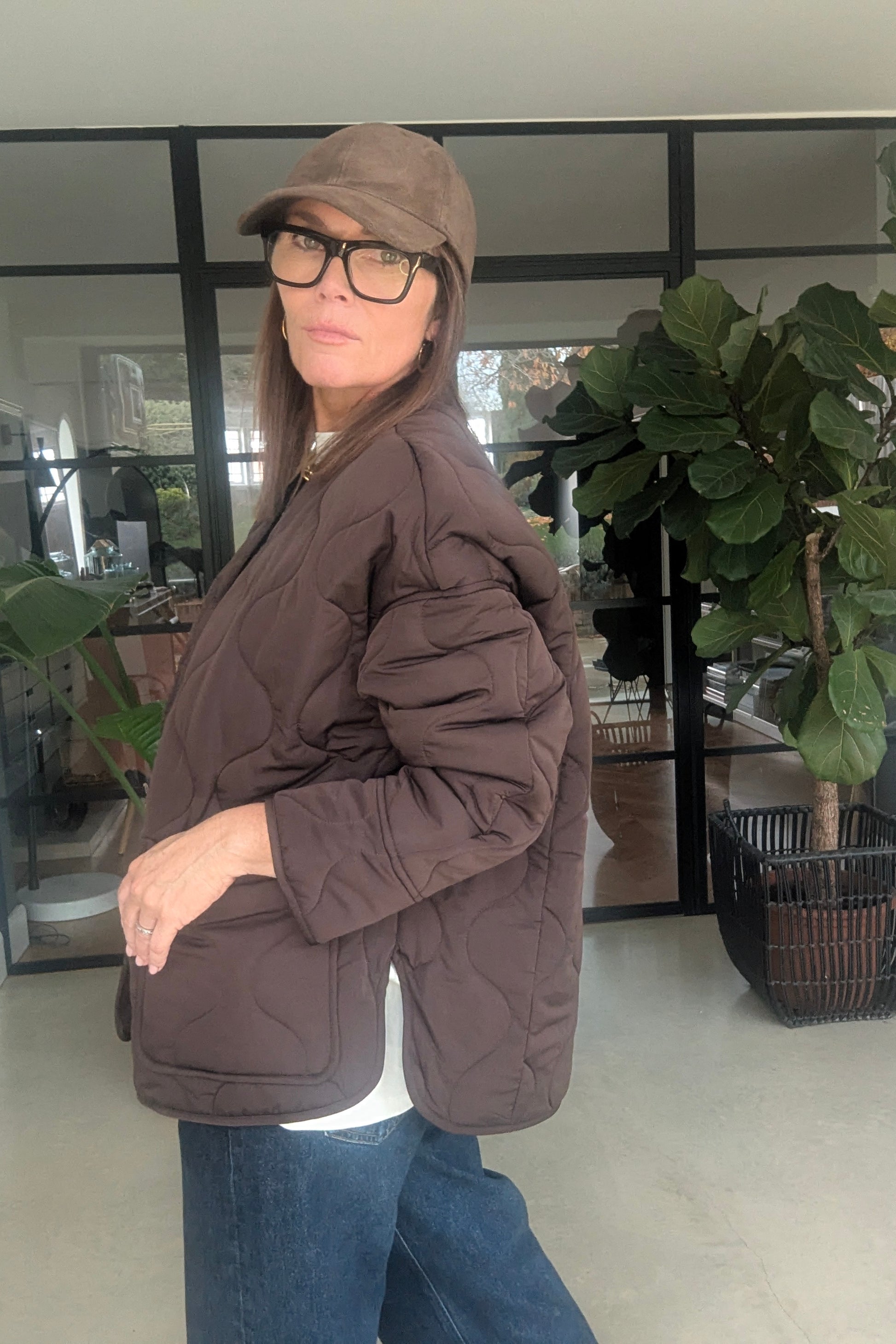 Person wearing a brown quilted jacket and cap indoors with plants in the background