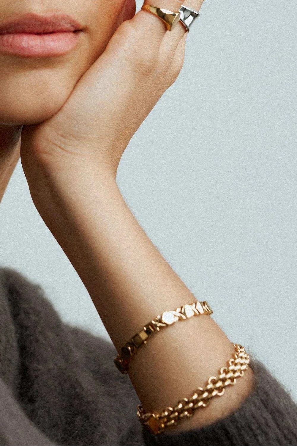 A close-up of a person's wrist and hand wearing three gold chain bracelets, with a matching gold ring on the finger, set against a light gray background.
