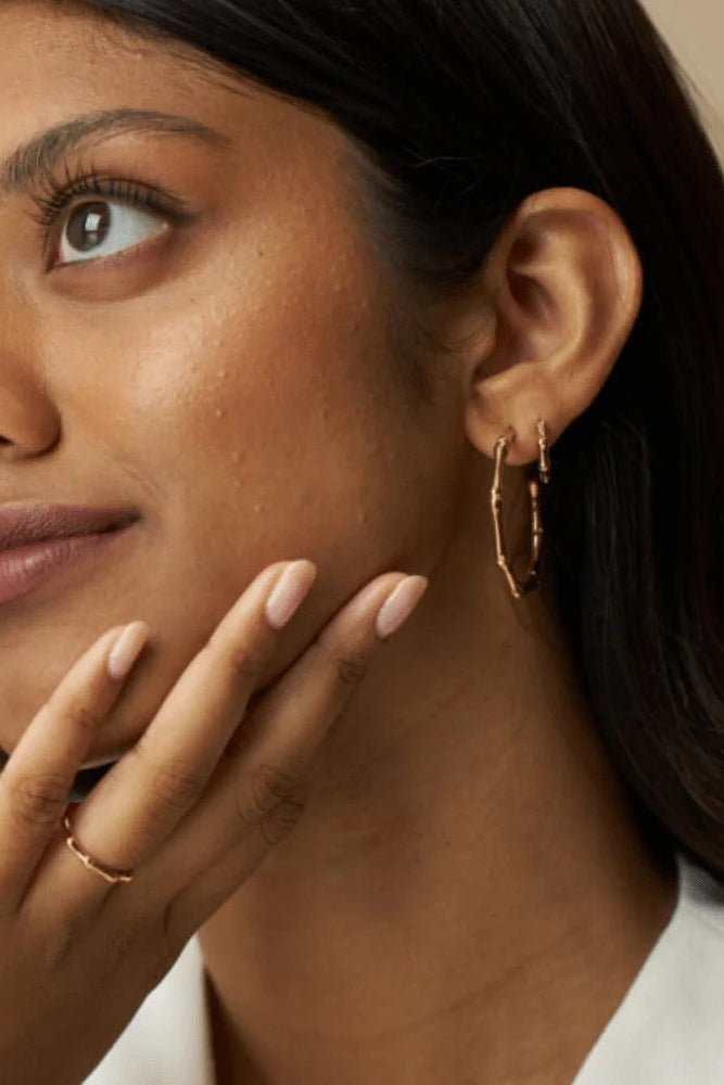Close-up of a woman wearing gold hoop earrings and a ring, with a neutral background.