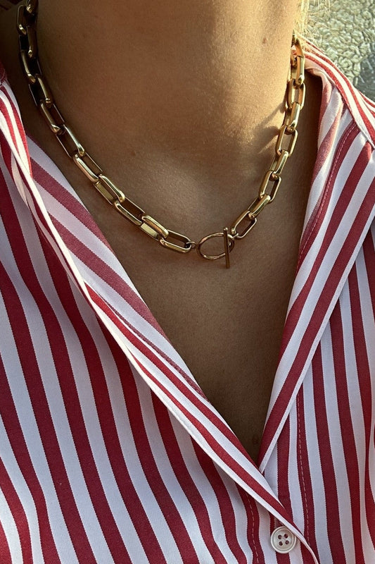A close-up of a person wearing a chunky gold chain necklace with a double-ring interlocking clasp, styled over a red and white striped collared shirt.