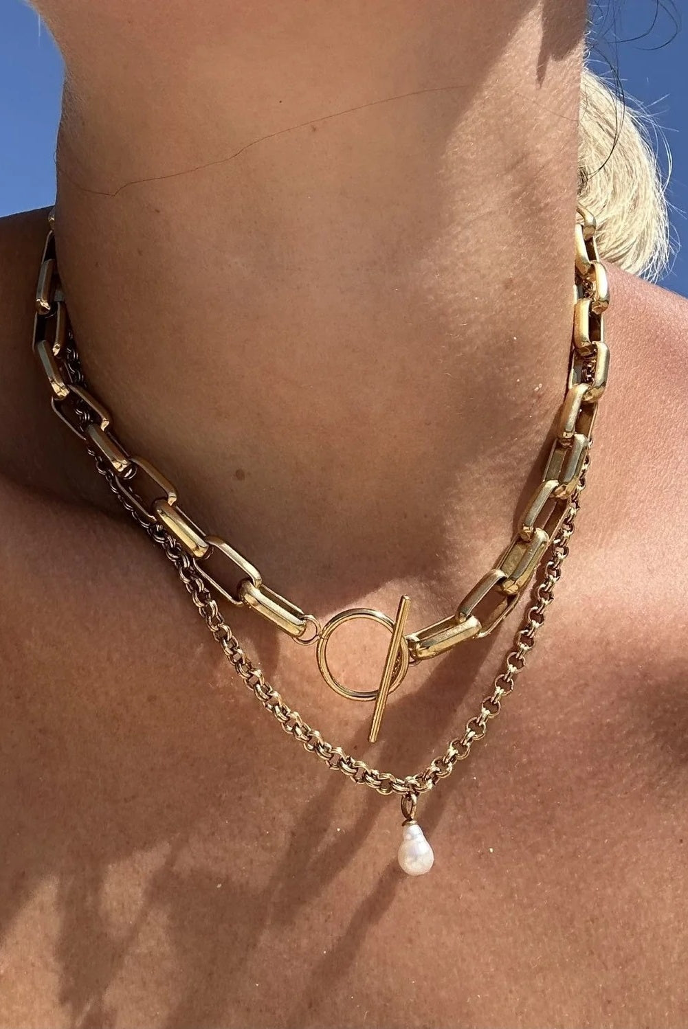 A close-up of a person's neck and upper chest wearing a layered gold-tone necklace. The necklace features a chunky link chain, a delicate chain with a small white teardrop pearl pendant, and a central circular ring with a bar detail.