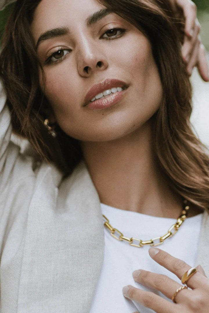 A close-up lifestyle shot of a woman wearing a chunky gold chain necklace, a gold ring, and a small gold hoop earring, paired with a white top and a light beige blazer.