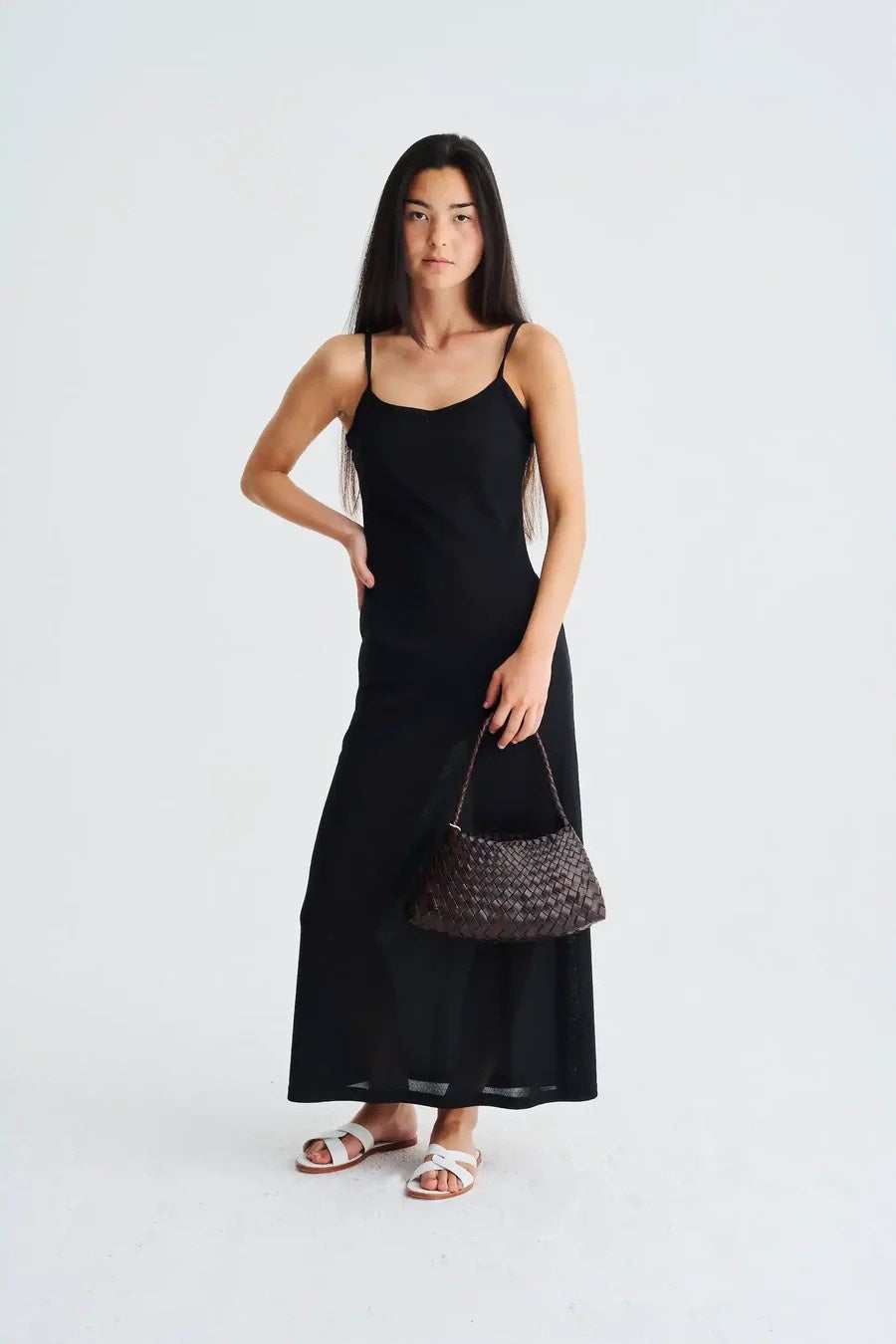 Woman wearing a black slip dress holding a brown handbag against a white background