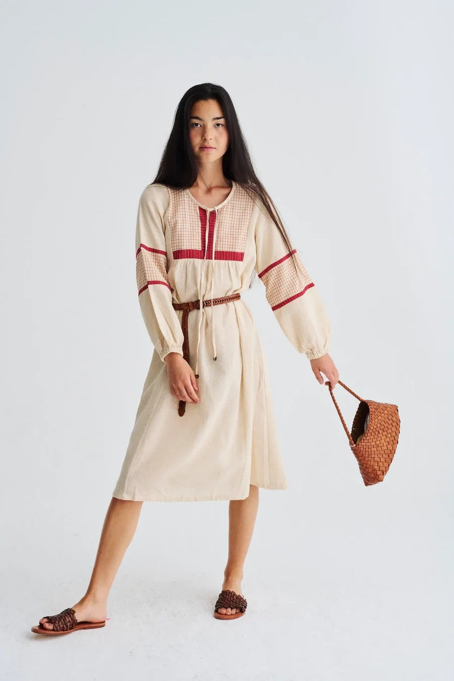 Woman wearing a beige dress with red accents, holding a woven bag on a white background