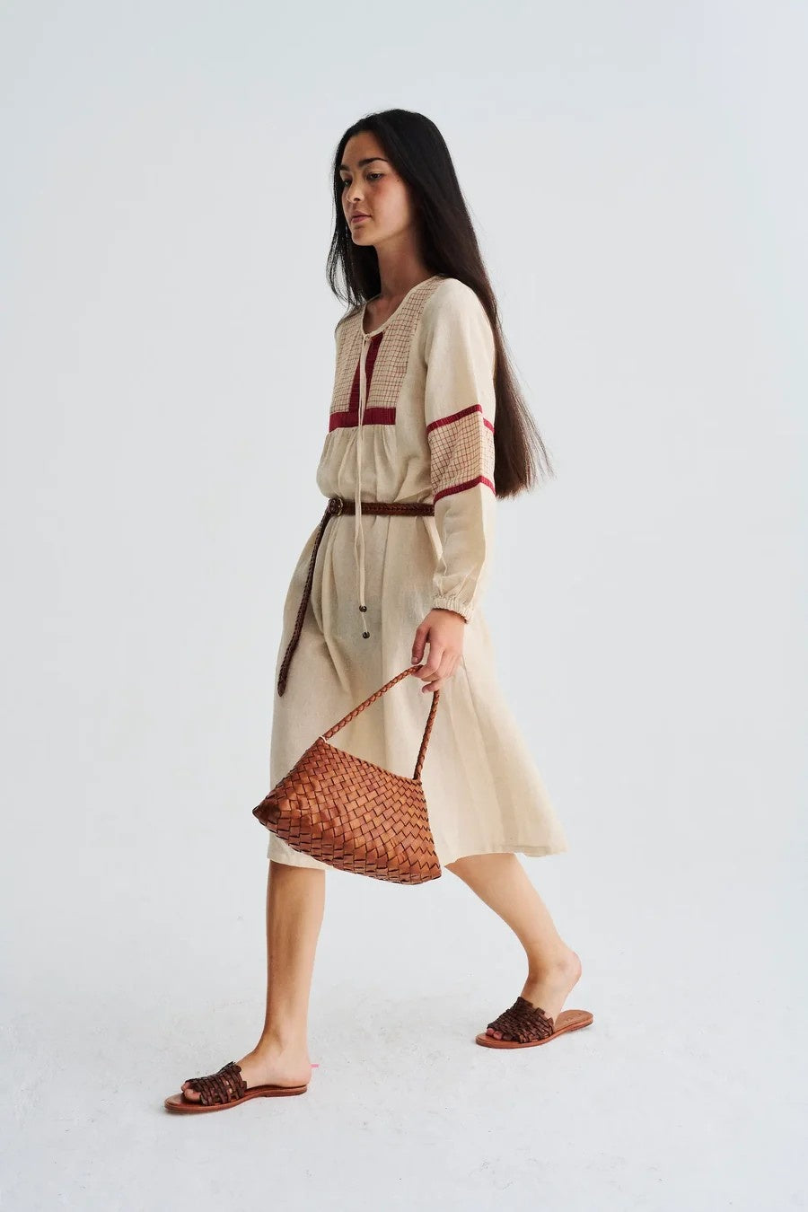 Woman in a beige dress with red accents holding a woven bag on a white background