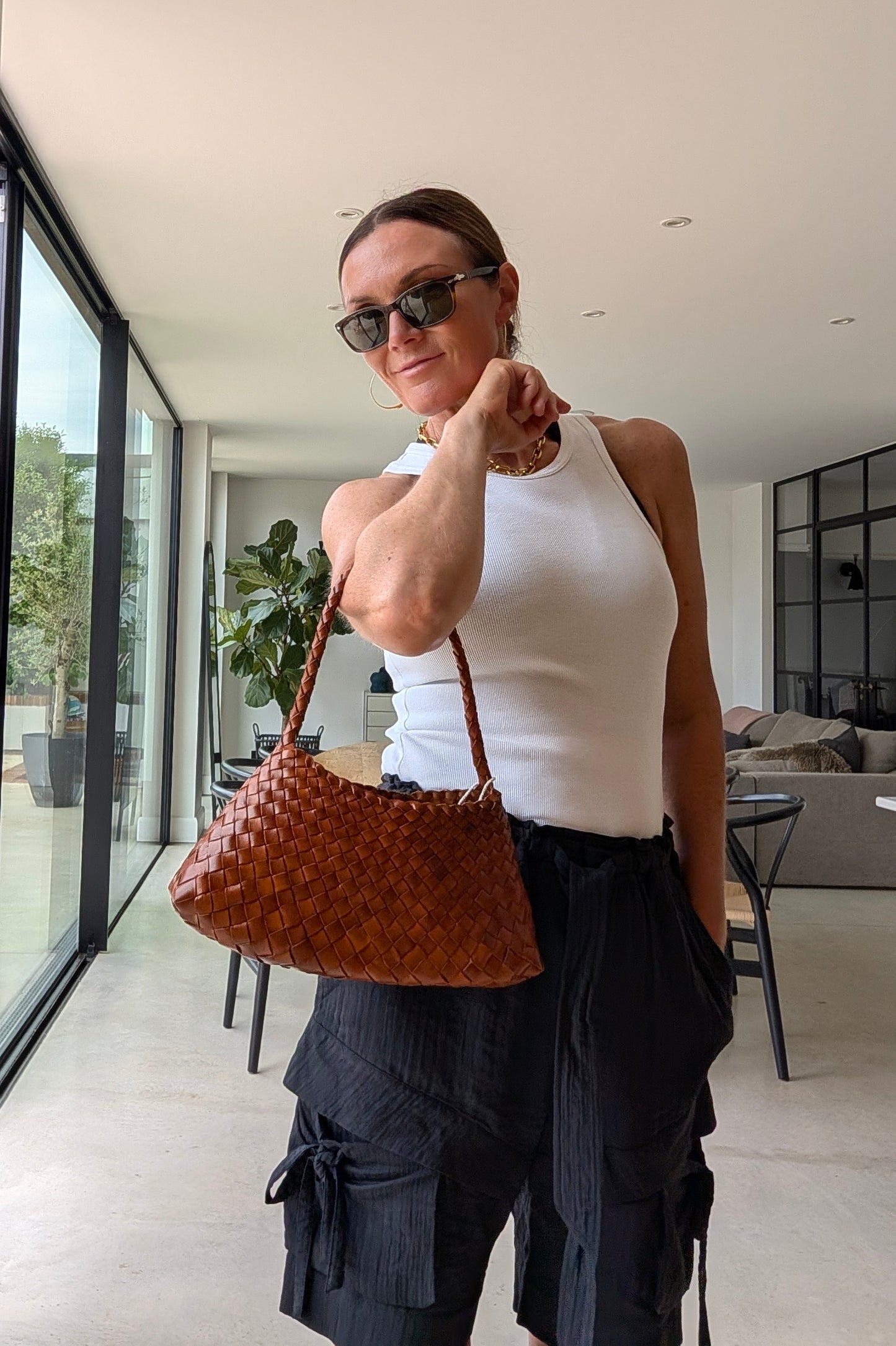 A woman wearing a white ribbed tank top and dark pleated cargo pants holds a brown woven leather shoulder bag in a modern indoor setting with large windows.