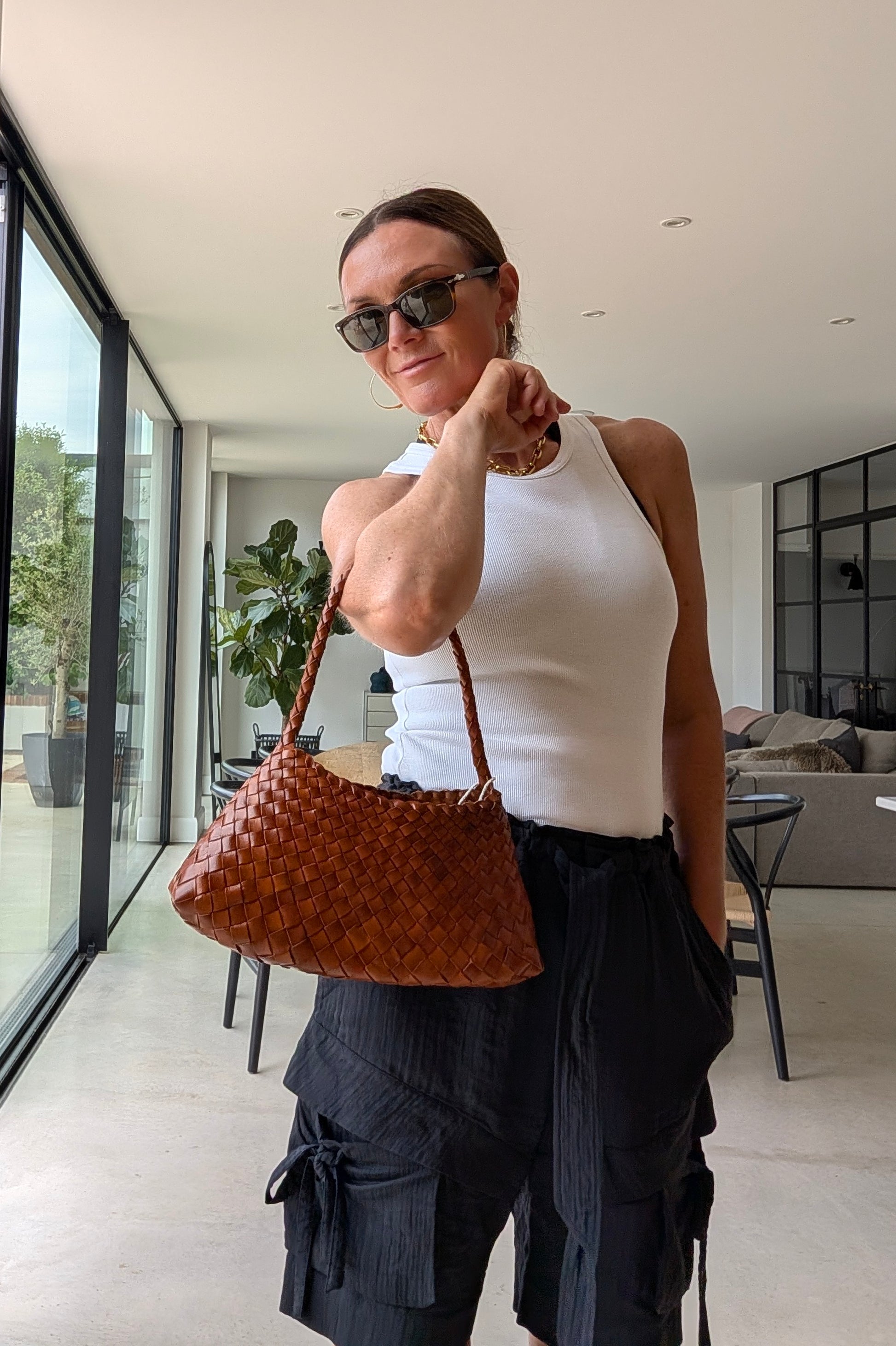 A woman wearing a white ribbed tank top and dark pleated cargo pants holds a brown woven leather shoulder bag in a modern indoor setting with large windows.