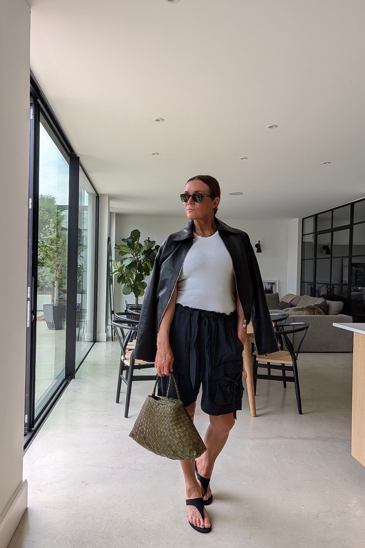 A woman walks through a modern indoor hallway wearing a black leather jacket, white t-shirt, black cargo shorts, black sandals, and holding a green woven handbag.