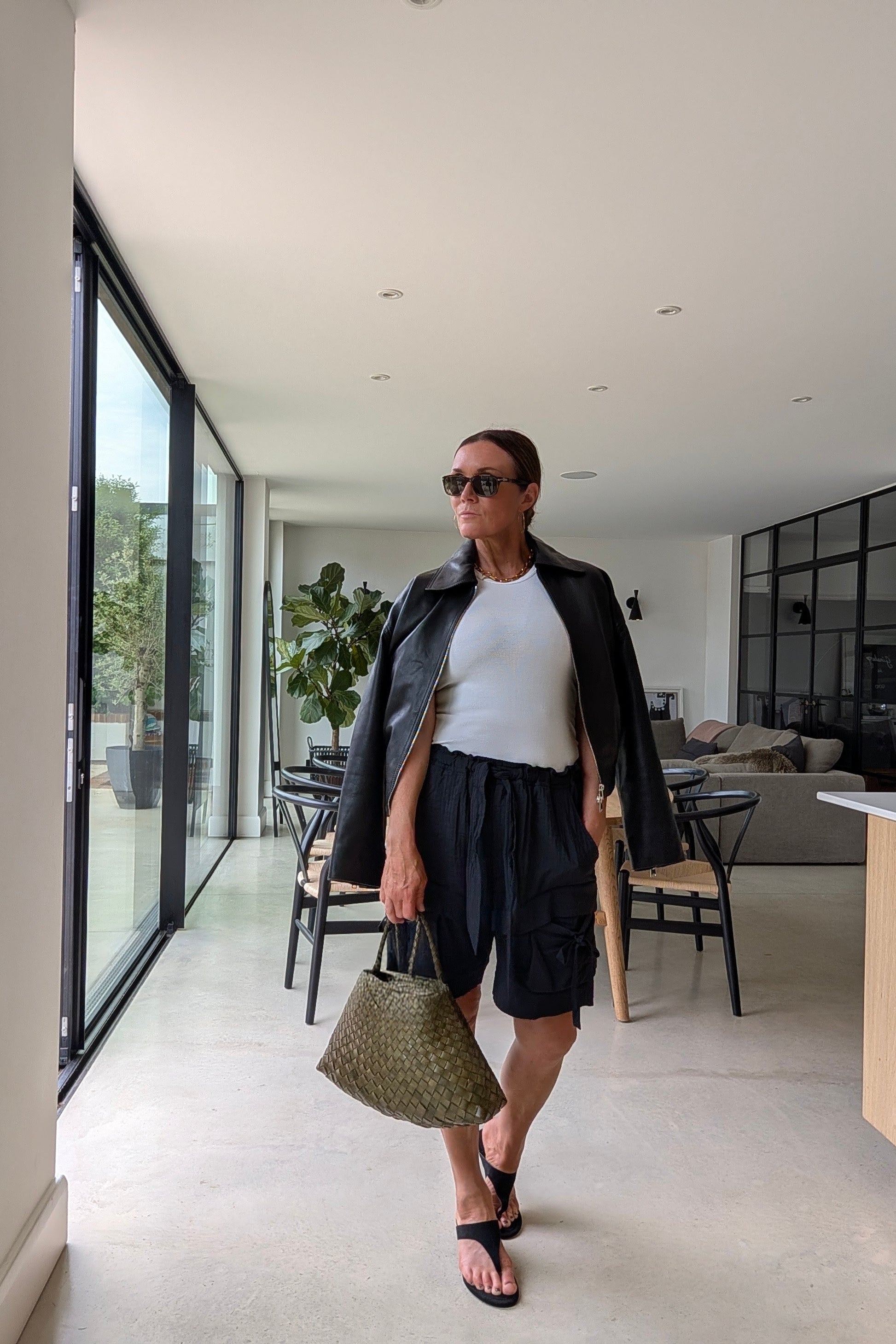 A woman walks through a modern indoor hallway wearing a black leather jacket, white t-shirt, black cargo shorts, black sandals, and holding a green woven handbag.