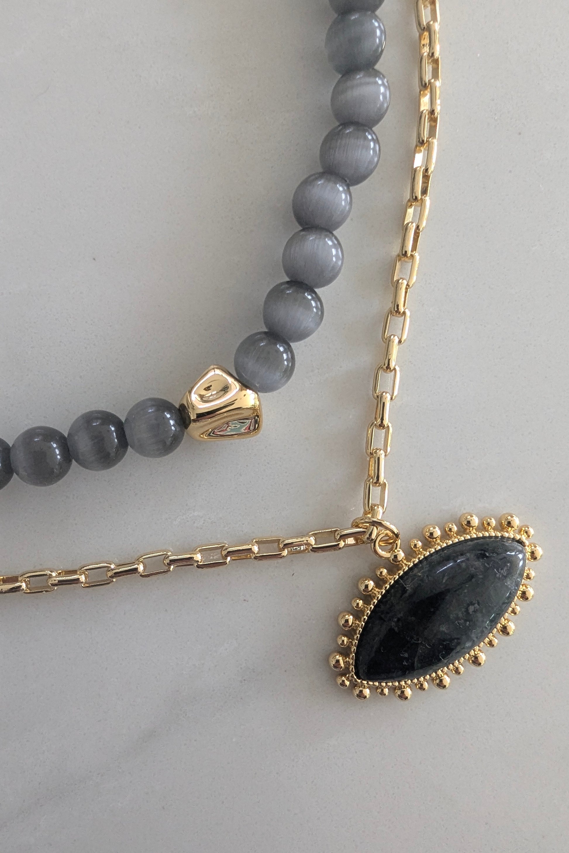 Two necklaces on a light gray background, one with gray beads and a gold clasp, the other with a black pendant and gold chain.