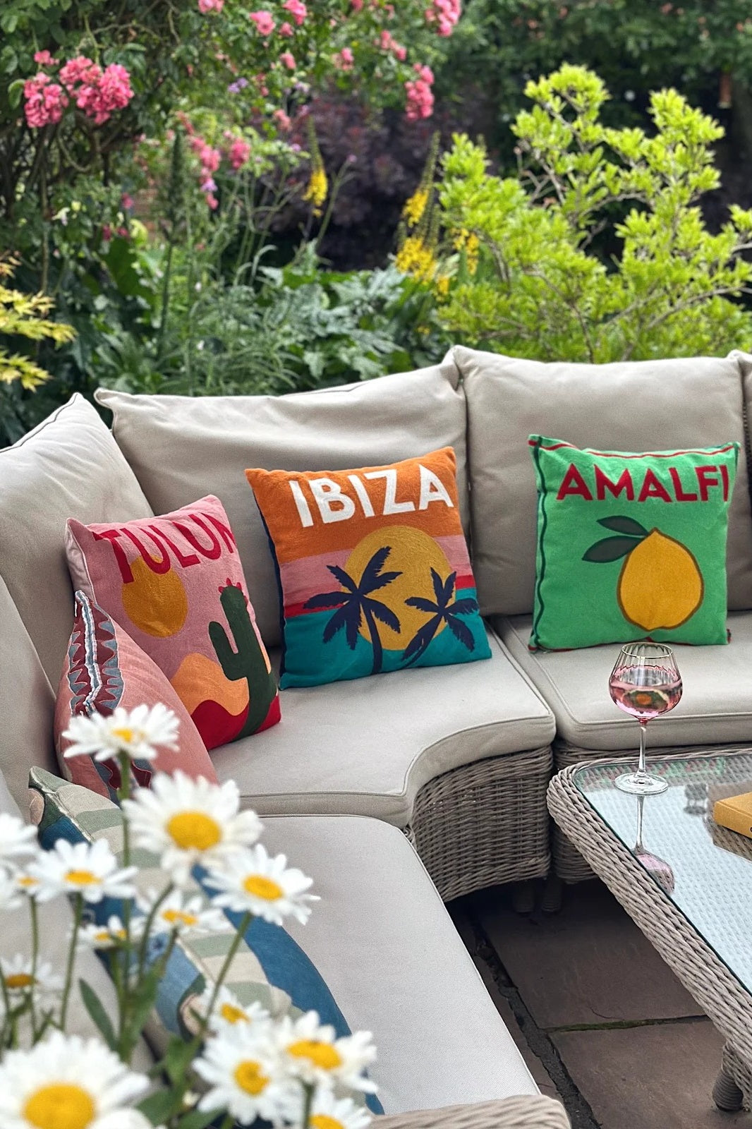 Outdoor sofa with colorful cushions labeled 'Tulum', 'Ibiza', and 'Amalfi' in a garden setting.