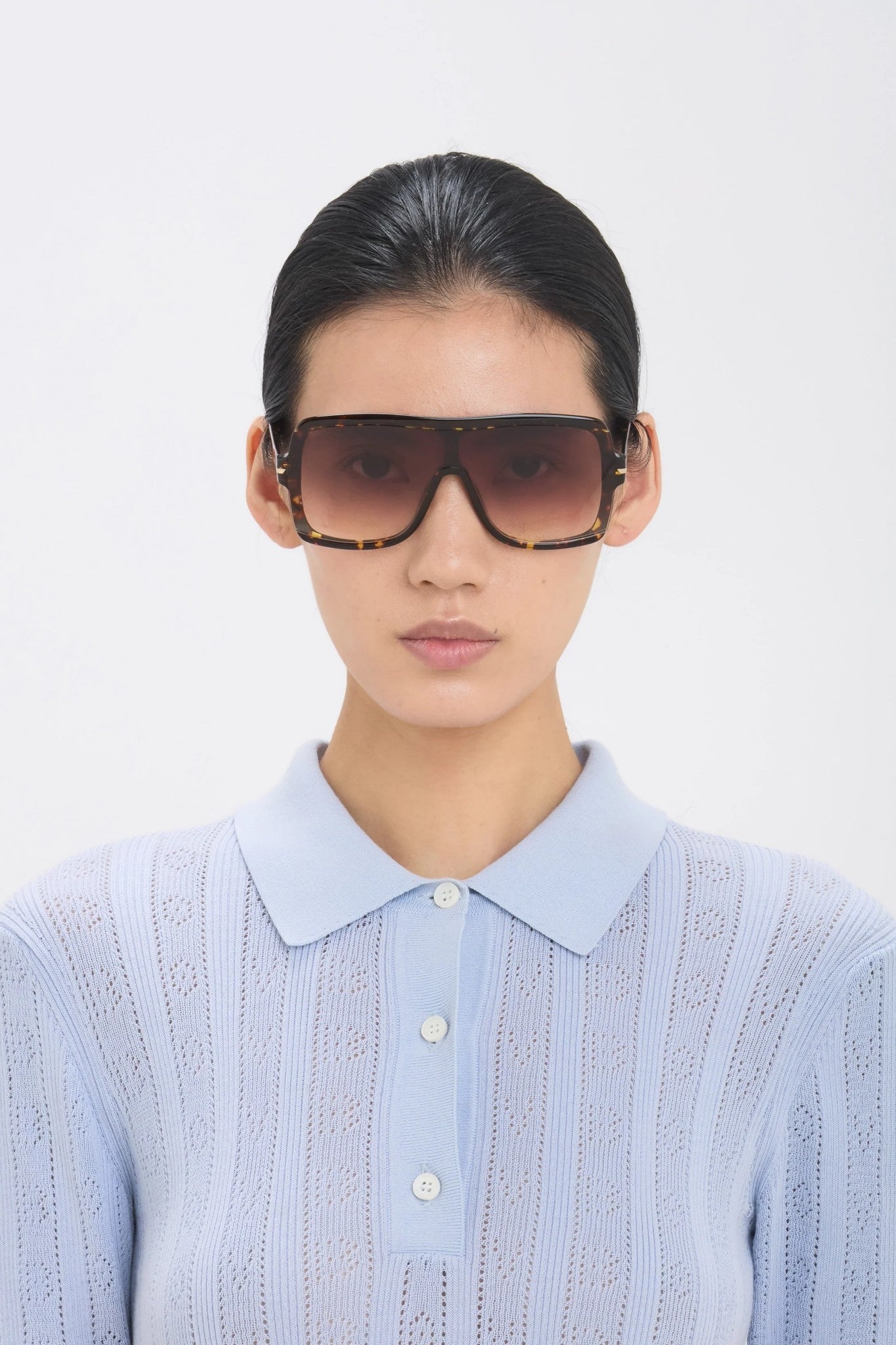 Person wearing a light blue knit sweater over a white shirt with sunglasses on a plain background
