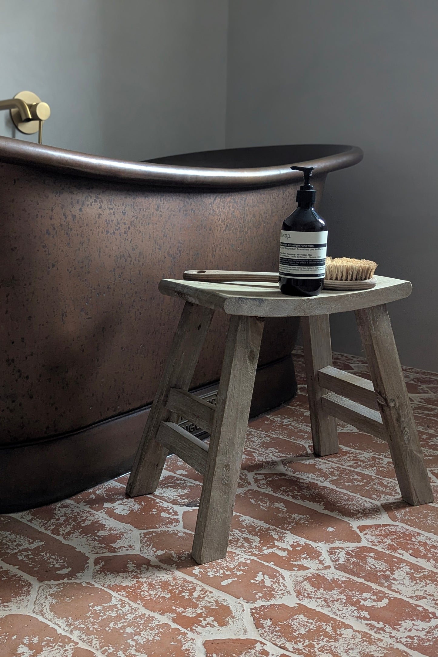 Wooden stool next to a freestanding bathtub with a bottle and brush on top, on a brick-patterned floor.