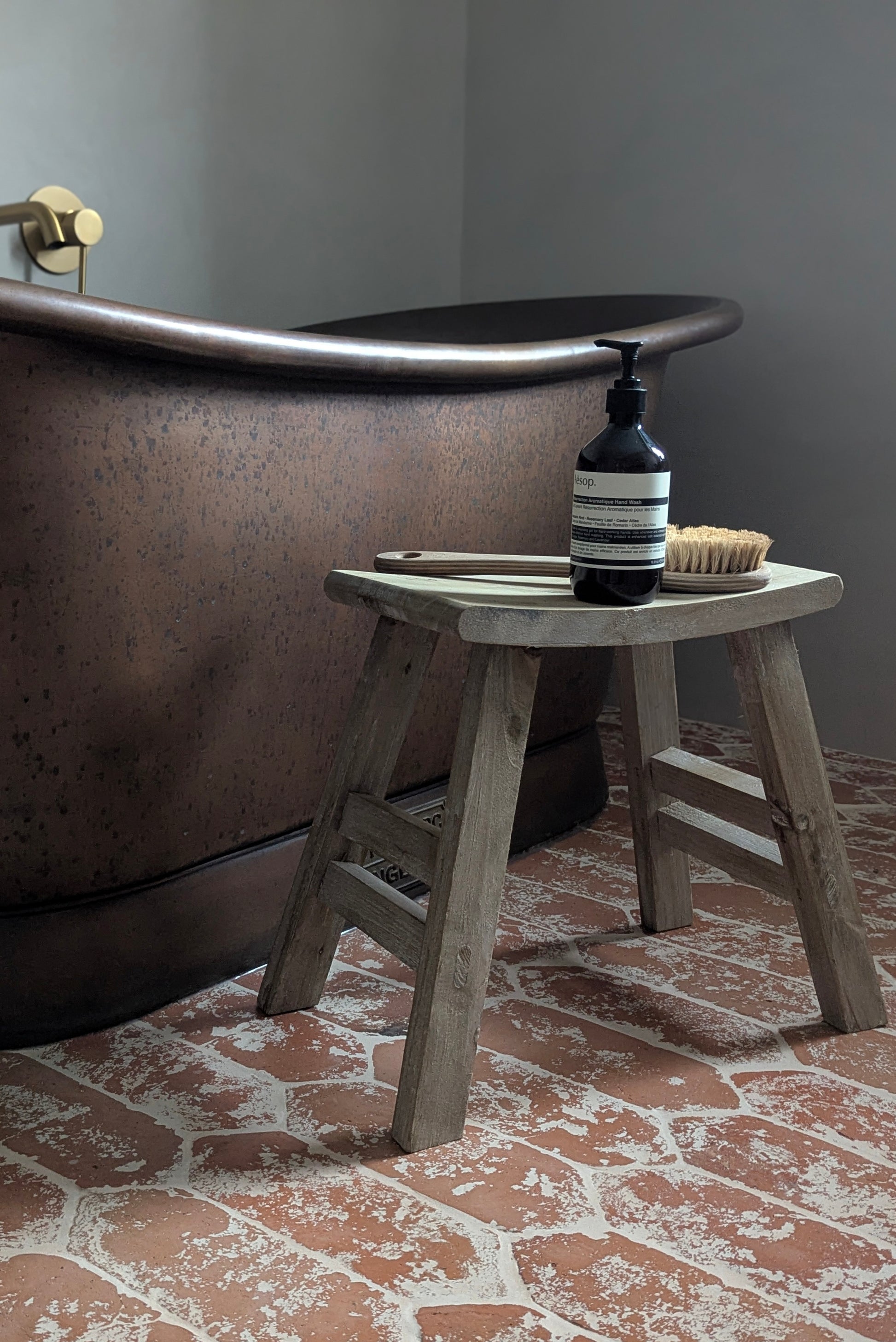 Wooden stool next to a freestanding bathtub with a bottle and brush on top, on a brick-patterned floor.