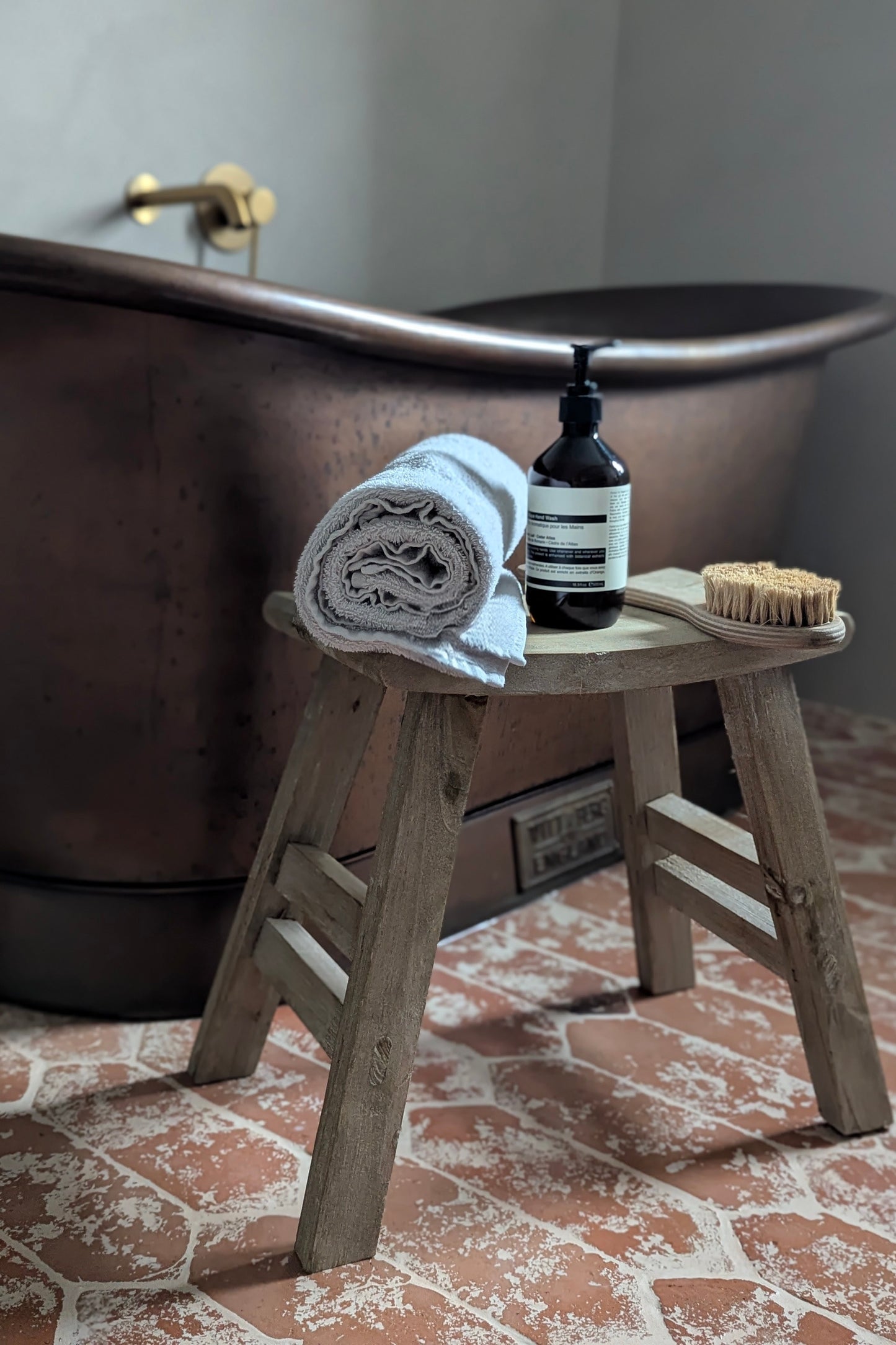 Bathroom setting with a wooden stool, bath towel, bottle, and brush in front of a freestanding bathtub.
