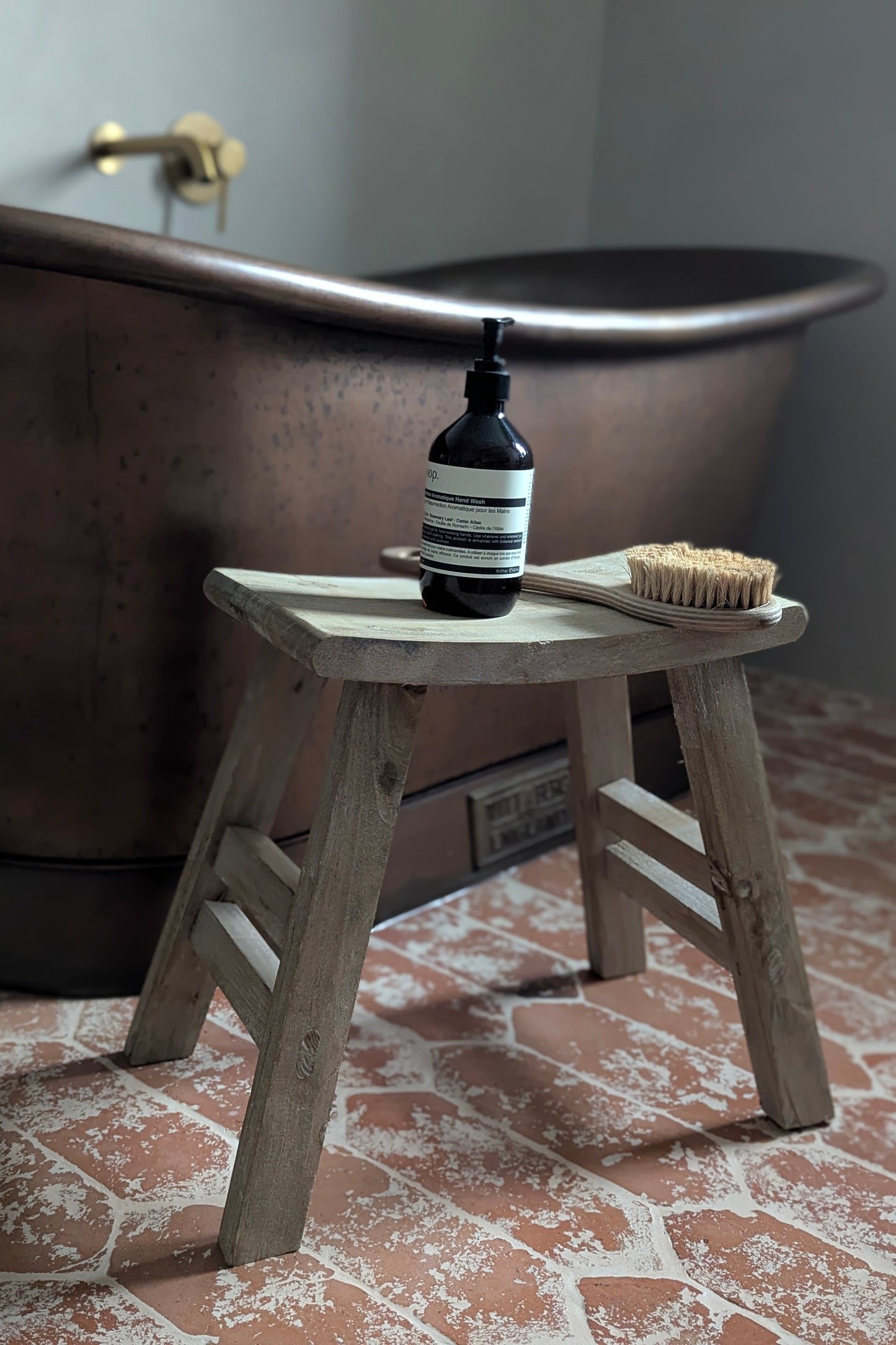Bottle of soap and brush on a wooden stool in front of a copper bathtub.
