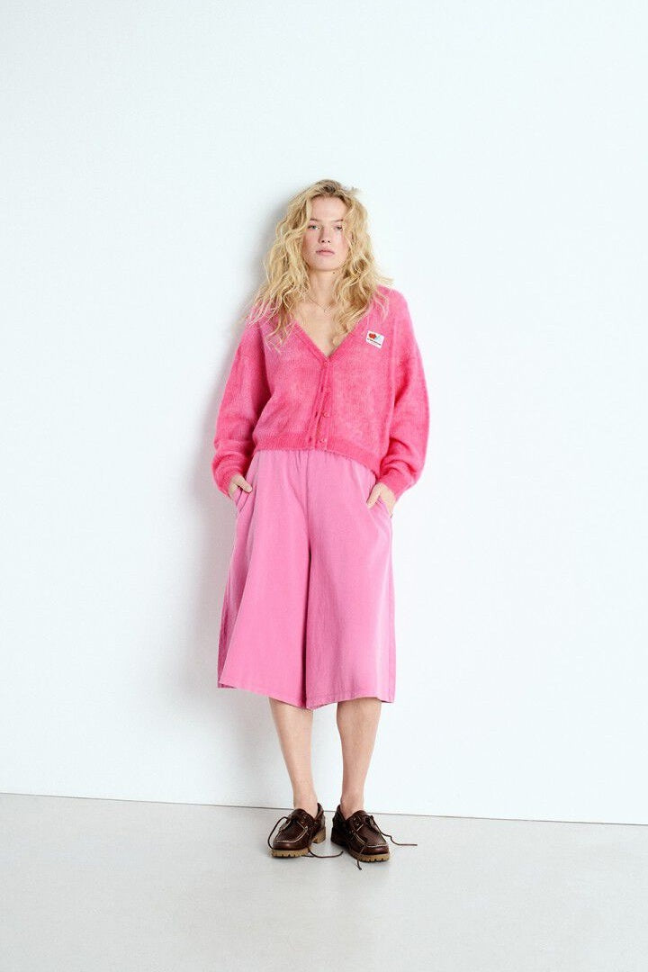 model wearing pink american vintage cardigan, pink skirt and brown deck shoes leaning against a white wall.